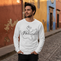 A man wearing a white long-sleeve t-shirt with a black map outline and the word 'Jalisco' printed on the front, standing on a cobblestone street in front of colorful buildings.