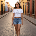 A woman walking down a cobblestone street wearing a white crop top with a black graphic print and light blue denim shorts.