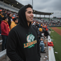 A man wearing a black hoodie with a 'LEONES' graphic stands in a stadium crowd, with bleachers, other spectators, and a field visible in the background.