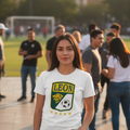 A woman standing outdoors in a sunny park-like setting wearing a white graphic t-shirt with a green and yellow 'LEON' soccer crest on the front, paired with black pants. The background shows a blurred crowd and sports field.