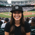 A smiling woman in a baseball stadium wearing a black cap with a white stylized logo and a black t-shirt, with the field and crowd blurred in the background.