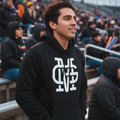 A man wearing a black hoodie with a large white stylized 'Guadalajara Unisexy Pullover Mariachis de Guadalajara' logo on the front, standing in stadium bleachers.