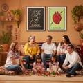 A family of six sits together on a woven rug in a warmly decorated room, surrounded by plants, shelves, and colorful toys. Two framed wall art prints hang above them, one black with a white anatomical heart illustration and one light green with a red anatomical heart illustration.