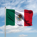 The Mexico national flag, featuring vertical stripes in green, white, and red colors with the national coat of arms, flying in the wind against a blue sky with clouds.
