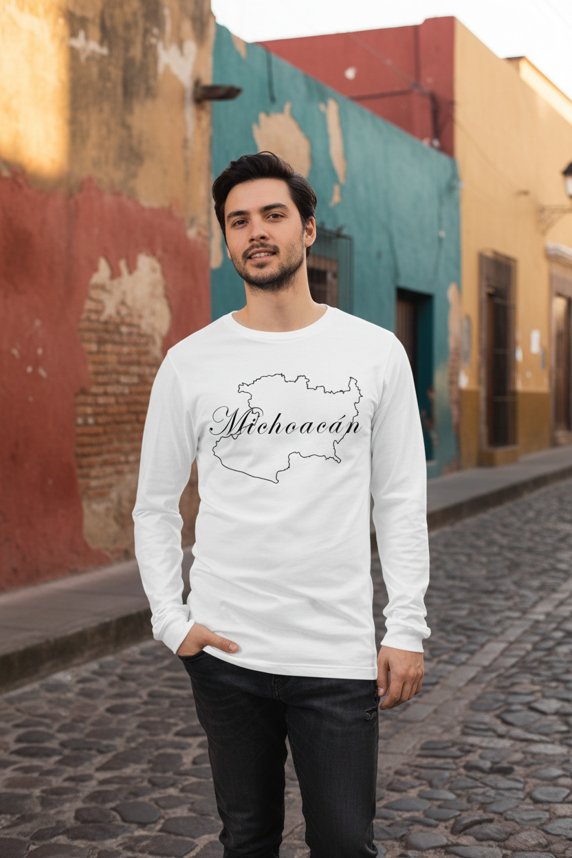 A man wearing a white long-sleeve t-shirt with a black outline map of Michigan and the word 'Michoacan' printed on the front, standing on a cobblestone street in front of colorful buildings.