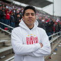 A man wearing a white pullover hoodie with a red and white Neckerés FC soccer crest stands with arms crossed in a stadium setting.