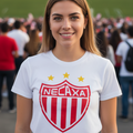 A smiling young woman stands outdoors wearing a white t-shirt with a red and white striped shield crest and the word 'Necaxa' across the chest, with a blurred crowd and green field in the background.