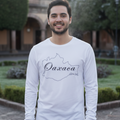 A smiling man wearing a white long-sleeve t-shirt with a black outline map of Oaxaca and the text 'Oaxaca' and 'Guatemala' printed on the front, standing outdoors in front of a historic building and green hedges.