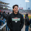 A man smiling in a stadium wearing a black hoodie with a 'Omecas' graphic on the front, paired with blue jeans.