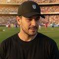 A man wearing a black baseball cap with a white crest Pachuca FC logo and a black t-shirt, standing in front of a blurred soccer stadium background.