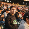 A man wearing a black Pericos de Puebla hoodie sitting in a crowded baseball stadium, smiling during a game.
