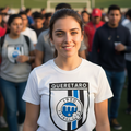 A smiling young woman stands on a soccer field wearing a white Queretaro FC athletic t-shirt with a black and blue crest graphic. The background shows a blurred crowd of people on the field.