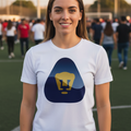 A smiling young woman stands on a sports field wearing a white t-shirt with a navy blue and yellow graphic of Pumas UNAM.