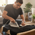 A man kneels beside a wooden table in a bright living room, ironing a black T-shirt laid flat. The shirt has a small colorful logo near the lower right area.