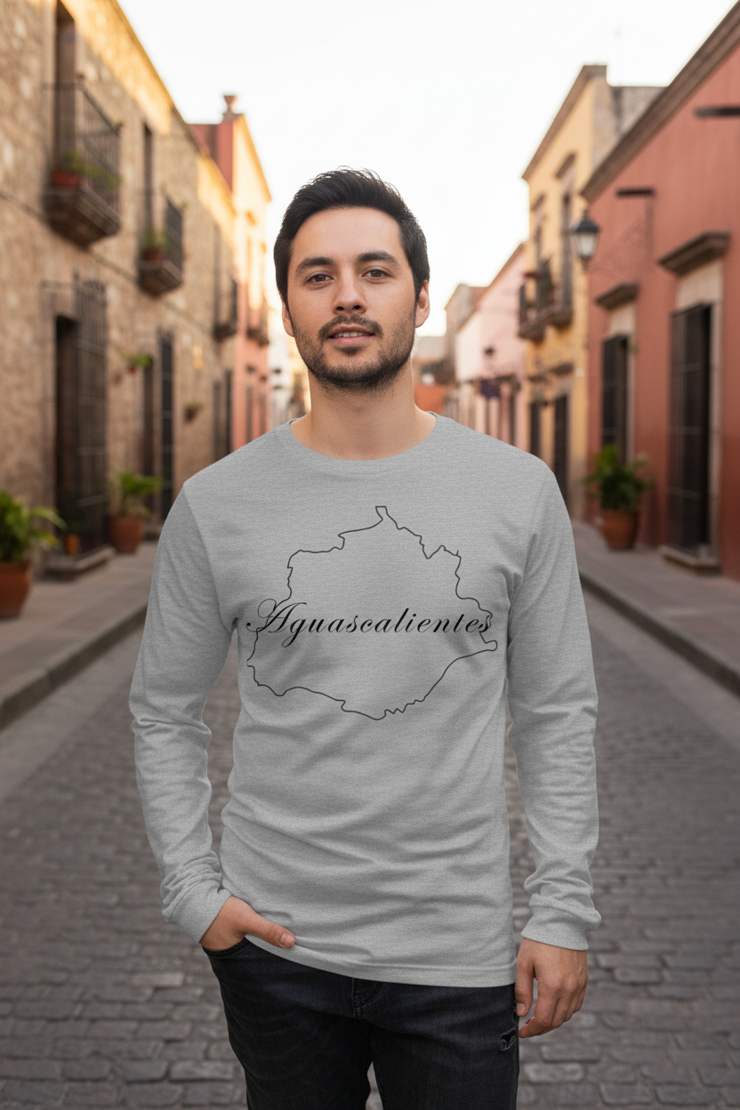 A man wearing a light gray long-sleeve t-shirt with a black outline map of the Aguascalientes state and the text 'Aguascalientes' across the chest, standing in a cobblestone street.