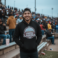 A man wearing a black hoodie with a 'Algodoneros de Union Laguna' graphic stands in a crowded outdoor stadium, with bleachers and other spectators blurred in the background.