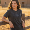 A woman wearing a dark gray mineral-wash t-shirt with a white outline logo on the chest, leaning against a wooden fence in a dry, golden field with adobe buildings in the background.