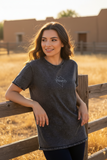 A woman wearing a dark gray mineral-wash t-shirt with a white outline logo on the chest, leaning against a wooden fence in a dry, golden field with adobe buildings in the background.