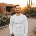 A man wearing a white long-sleeve t-shirt with a black outline of the state of Chihuahua and the word 'Chihuahua' written across it, standing outdoors in a desert setting at sunset.