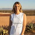 A smiling woman stands outdoors in a desert landscape wearing a white sleeveless top with a black outline of Chihuahua and the word 'Chihuahua' printed across the front, paired with black pants.