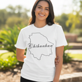 A woman wearing a white short-sleeve t-shirt with a black outline of the state of Chihuahua and the word 'Chihuahua' written in script across it, standing outdoors in a sunny garden setting.