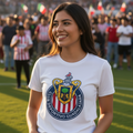 A woman wearing a white short-sleeve t-shirt with the Guadalajara Chivas soccer team crest printed on the front, standing on a grassy field with a stadium crowd in the background.