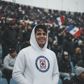 A smiling man in a white hoodie stands in a crowded stadium, with a blue and white Sporting Cruz Azul crest printed on the front of the sweatshirt.