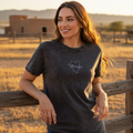 A woman wearing a dark charcoal gray t-shirt with a white outline graphic of the state of Texas and the word 'Dakotas' inside, leaning on a wooden fence in a dry grass field at sunset.