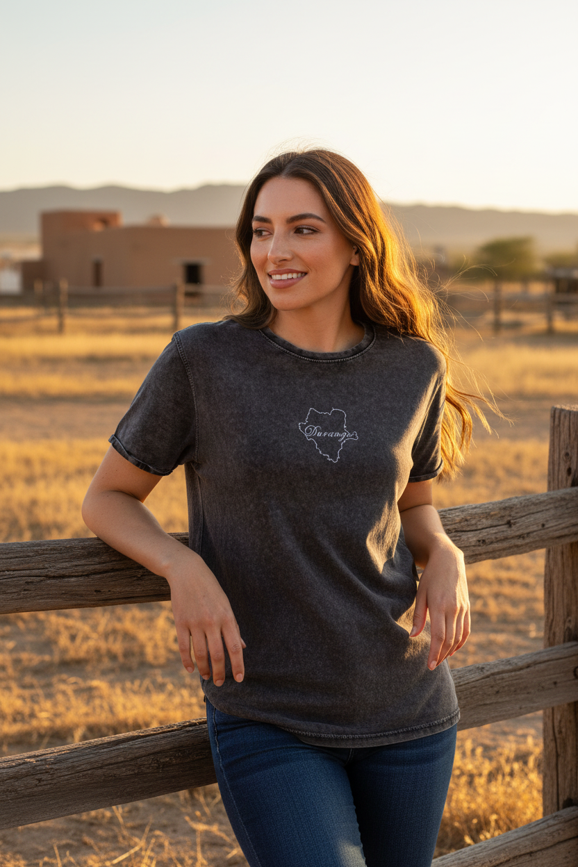 A woman wearing a dark charcoal gray t-shirt with a white outline graphic of the state of Texas and the word 'Dakotas' inside, leaning on a wooden fence in a dry grass field at sunset.