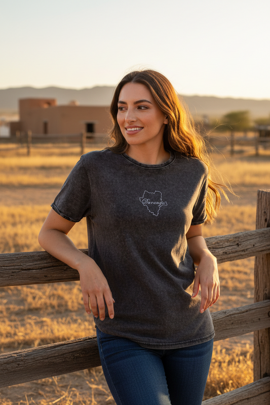 A woman wearing a dark charcoal gray t-shirt with a white outline graphic of the state of Texas and the word 'Dakotas' inside, leaning on a wooden fence in a dry grass field at sunset.