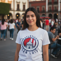 A woman wearing a white short-sleeve t-shirt with a red and blue Atlético San Luis logo, standing in a public square with blurred buildings and people in the background.