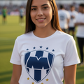 A smiling young woman standing on a soccer field wearing a white Monterrey FC t-shirt with a blue and white shield logo and gold stars.