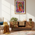 A framed colorful religious art print of Our Lady of Guadalupe hangs on a light beige wall above a wooden console table in a styled living room setting with a chair, throw blanket, plants, and a patterned rug.