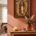 A framed religious painting of Our Lady of Guadalupe hangs on a terracotta-colored wall above a rustic wooden console table decorated with candles, a glass vase of dried grasses, and small decorative figurines.