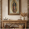 A framed religious print of Our Lady of Guadalupe hangs on a beige wall above a rustic wooden console table decorated with candles, books, a basket, and dried grass in a styled interior setting.