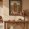 A framed religious painting of Our Lady of Guadalupe hangs on a warm beige plaster wall above a rustic wooden console table styled with candles, books, and a vase of dried grasses.