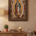 A framed religious art print of Our Lady of Guadalupe hanging on a beige wall above a wooden console table with a lamp, cactus, books, and a candle.
