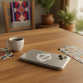 Silver smartphone on a wooden table with a die-cut bubble-free sticker shaped like the state of Aguascalientes — featuring white outline and cursive “Aguascalientes” text, surrounded by a coffee cup, colorful Mexican tiles, and warm natural light, creating an artisanal and cozy aesthetic that highlights regional pride and craftsmanship.