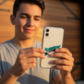 A young man smiling while holding up a white smartphone to show a close-up of a green and red 'Bravos de Leon' graphic decal on the back of the phone.