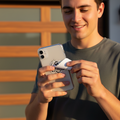 A smiling young man in a dark gray t-shirt holds a silver smartphone and a small white rectangular accessory with a circular Guerreros logo, standing outdoors in front of a modern wooden-paneled building.