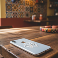  Silver smartphone placed on a wooden café table with a die-cut bubble-free sticker reading “Hidalgo” in cursive — surrounded by warm lighting, patterned tiles, and rustic décor, symbolizing Mexican regional pride and artisanal design.