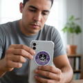 Man seated at a wooden table holding a smartphone with a clear case featuring a die-cut bubble-free sticker of the Mazatlán F.C. logo — a circular purple design with a white border, anchor, and shield emblem representing the Mexican football club.