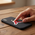 A black phone case with a red and white stylized 'A' logo, shown on a smartphone resting on a wooden table while a hand touches the case.
