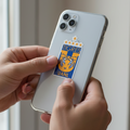 Close-up of hands holding a smartphone with a die-cut bubble-free sticker of the Tigres UANL logo — featuring a gold tiger head on a blue background with four stars above, representing the Mexican football club’s identity and pride.
