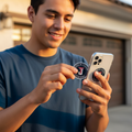 A smiling young man outdoors in front of a garage holds a smartphone with a circular Toros de Tijuana, white, and he is also holding a matching round sticker or patch in his other hand.
