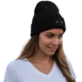 A woman wearing a black knit beanie with a small white embroidered Aguascalientes logo on the front, standing outdoors in front of a warm-toned building with a dark wooden door.