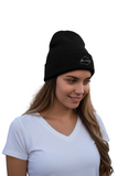 A woman wearing a black knit beanie with a small white embroidered Aguascalientes logo on the front, standing outdoors in front of a warm-toned building with a dark wooden door.