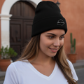 A woman wearing a black knit beanie with a small white embroidered Aguascalientes logo on the front, standing outdoors in front of a warm-toned building with a dark wooden door.