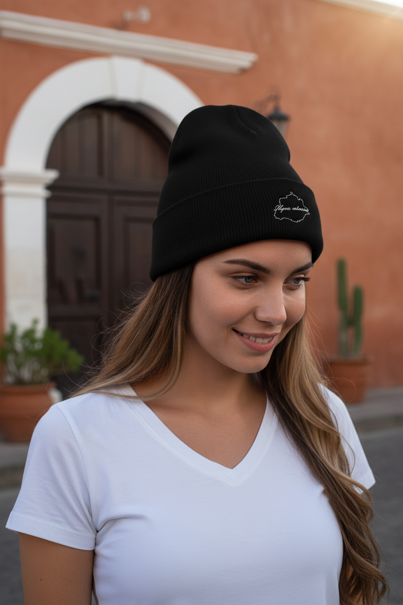 A woman wearing a black knit beanie with a small white embroidered Aguascalientes logo on the front, standing outdoors in front of a warm-toned building with a dark wooden door.