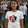 A woman standing on a sports field wearing a white t-shirt with an Atlas FC red, black, and white shield graphic on the chest, with a blurred crowd in the background.